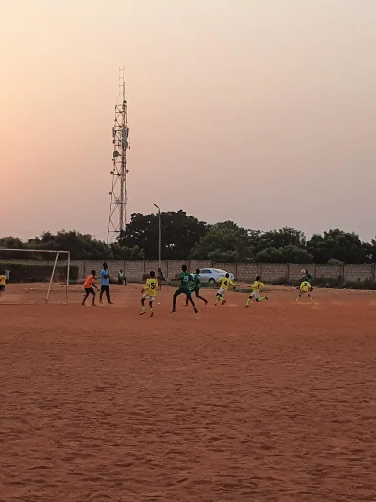 Young players in yellow and green kits playing football on a dirt pitch in Ghana at dusk