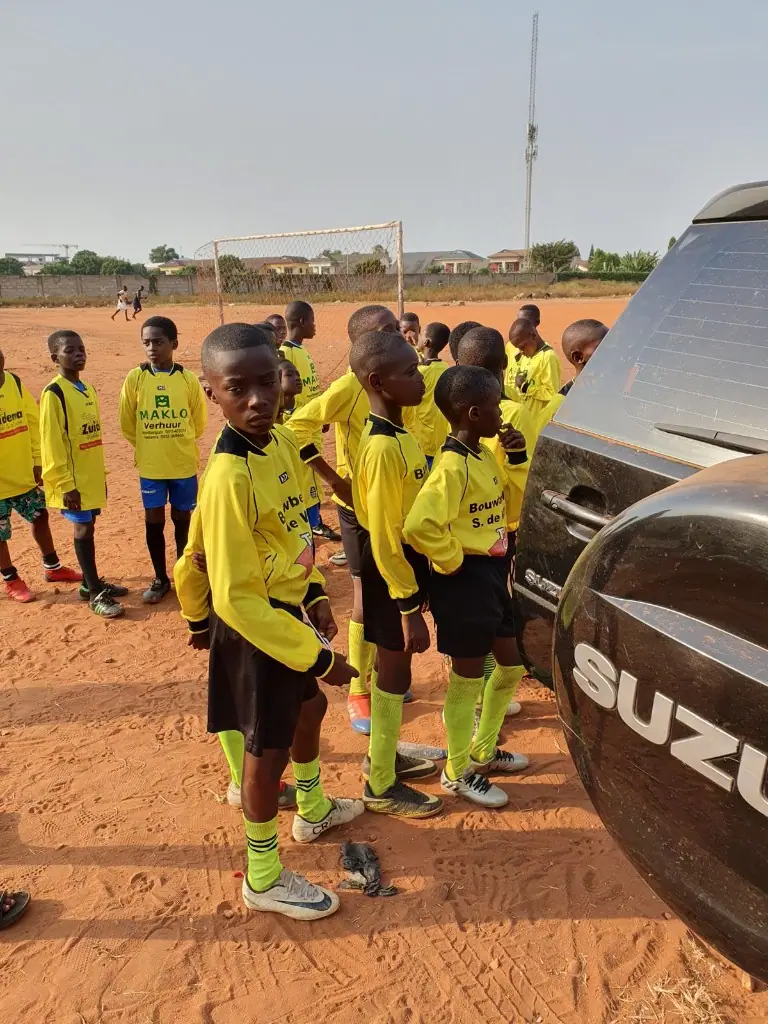 A youth football squad in yellow kits standing together on a dirt pitch in Ghana