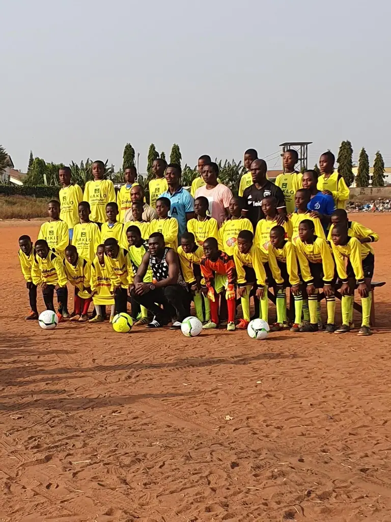 A large youth football team in yellow kits posing with coaches on a dirt pitch in Ghana